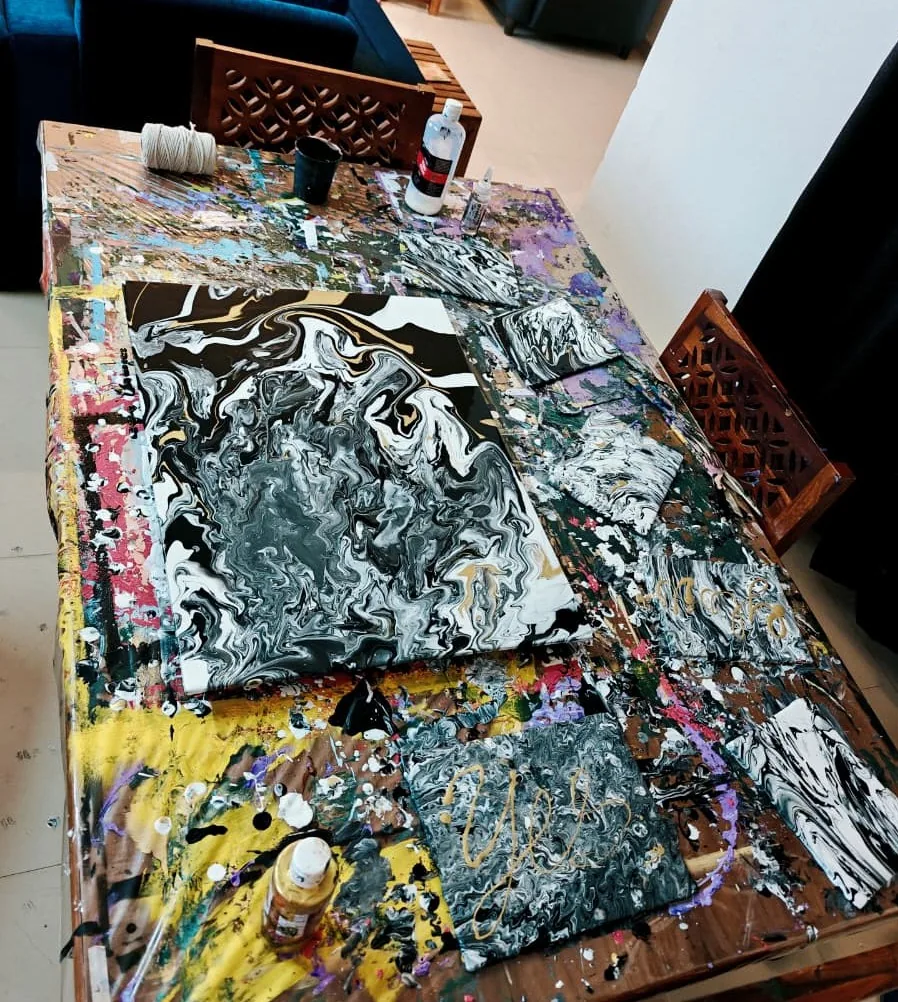 A photograph looking across a heavily paint-covered studio table on which a large black, white and gold marbled pour is drying, surrounded by several smaller canvases in the same monochrome palette, two of them inscribed with gold script, with bottles of paint and medium at the edges.