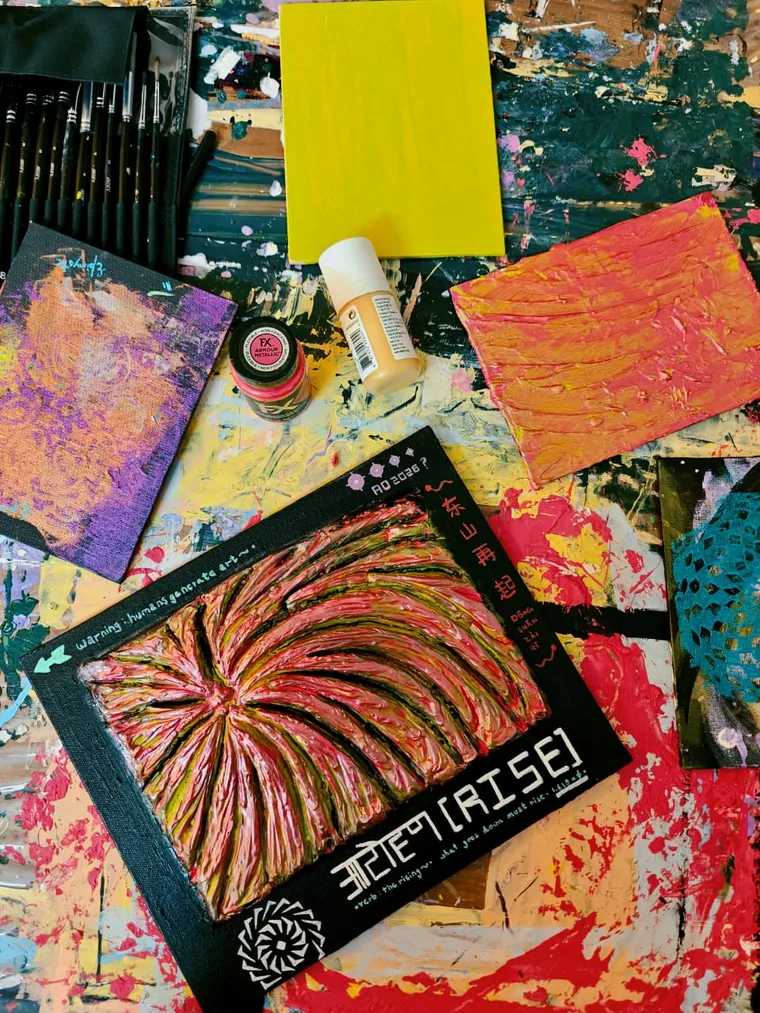 A photograph taken at an angle of the lettered relief painting lying on a paint-covered studio table among other freshly painted canvases, a bottle of medium and a jar of metallic paint, with brushes at the edge of the frame.