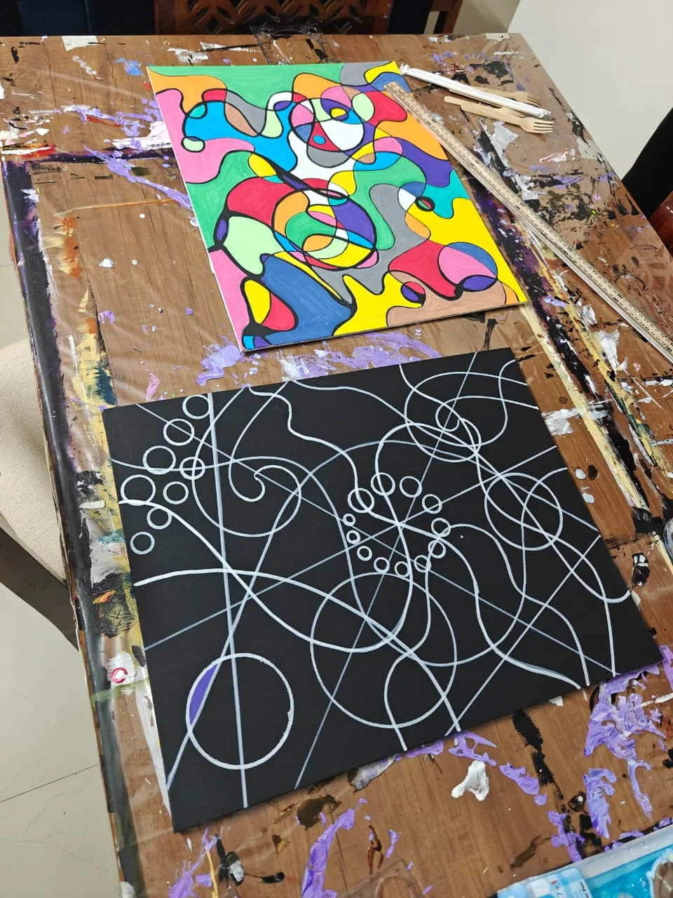 A photograph of two canvases on a paint-splattered wooden studio bench: above, a finished cloisonne painting of black-outlined cells filled with bright flat colour; below, a black canvas bearing only looping white lines and circles, the drawn stage before the colour is added.