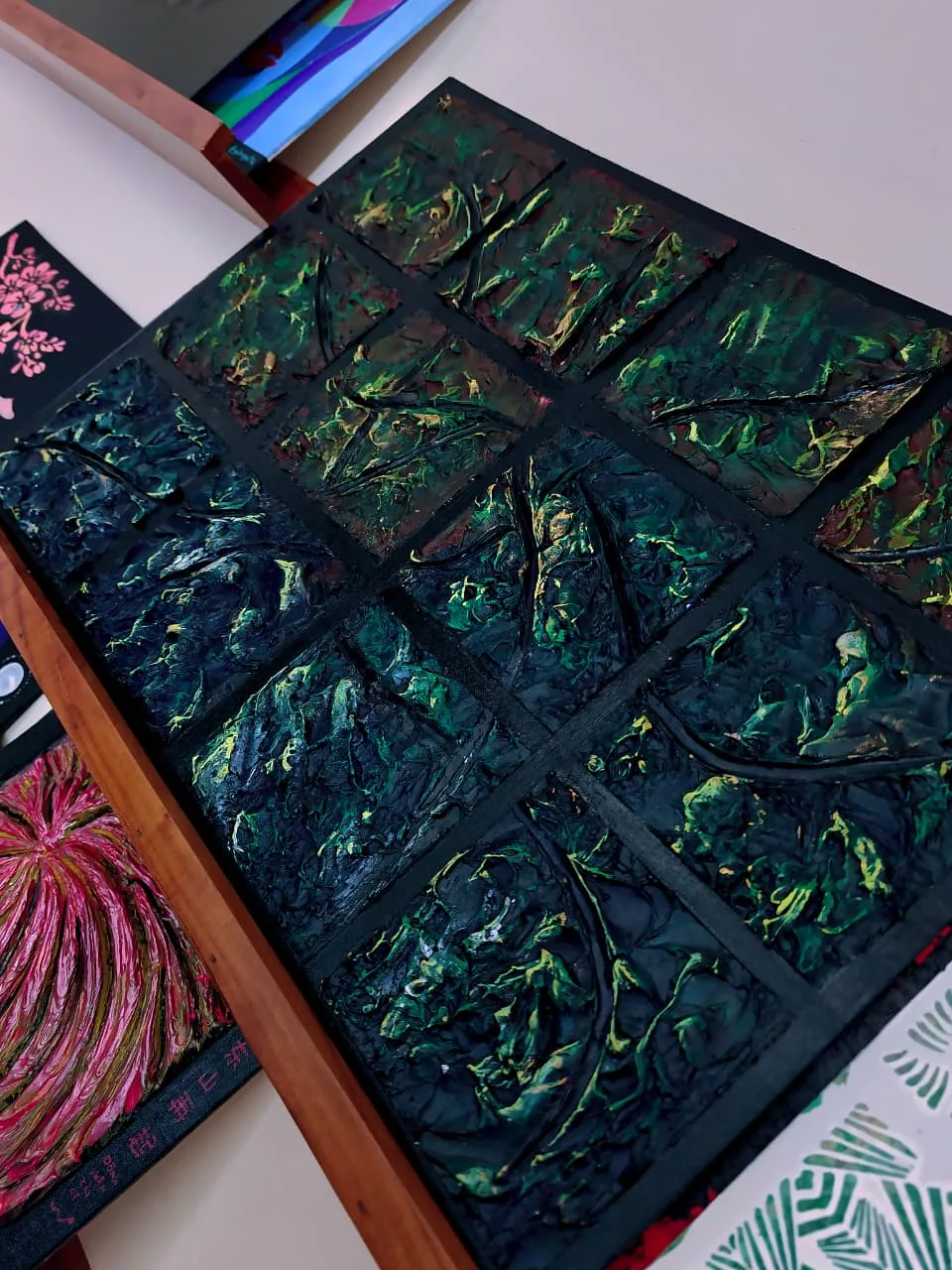 A relief painting divided into nine panes of dark green and black acrylic paste, seen at an angle on a studio table beside other canvases.