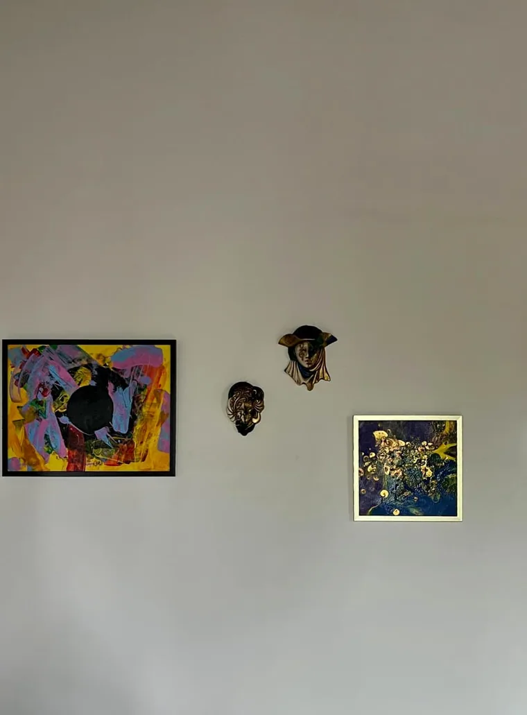 A photograph of a wall hung with two framed abstract paintings — one of a black horse on a bright expressive ground, one a dark blue-and-gold pour — flanking two sculptural face masks.