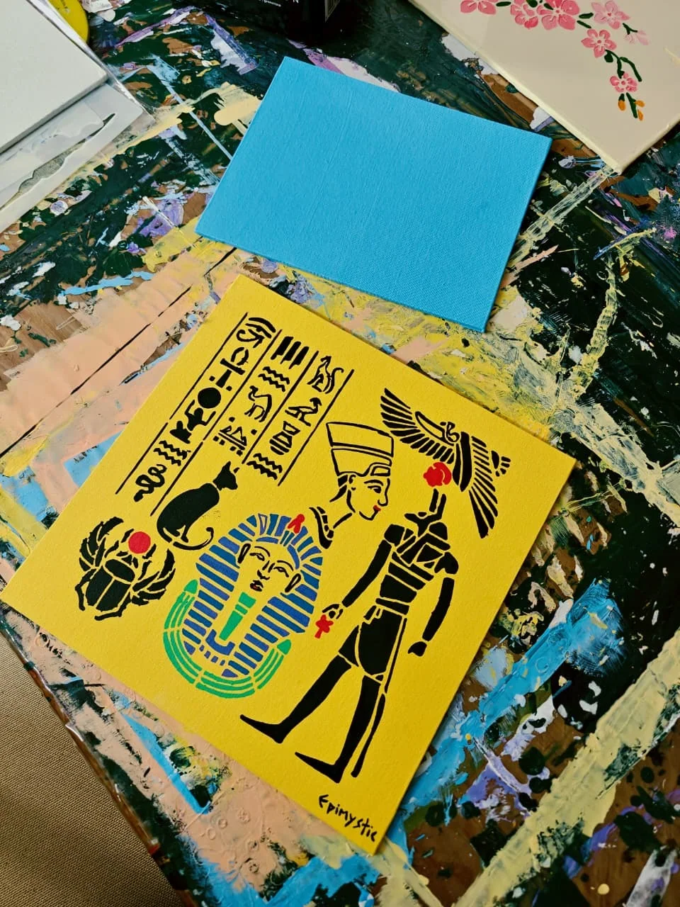 A flat graphic painting on a bright yellow ground: bands of black hieroglyphs, a scarab beetle with a red sun disc, a seated black cat, the blue-and-gold mask of Tutankhamun, Nefertiti in profile with her crown, and a winged figure holding an ankh.