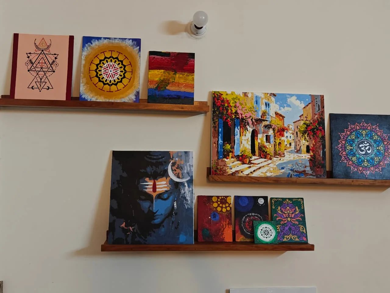 A photograph of a pale wall with three wooden picture-ledges holding many framed paintings: a blue meditating Shiva, a sunlit Mediterranean street scene, an Om mandala, sacred-geometry panels, sun-mandalas, and several small abstract and ornamental works.