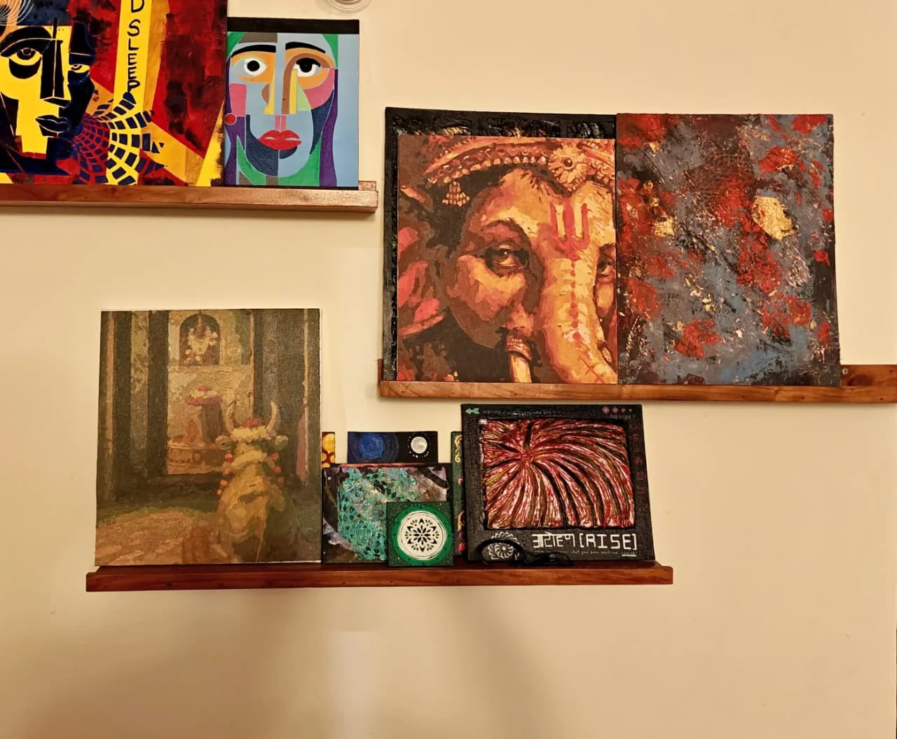 Three wooden picture ledges on a cream wall: on the top ledge two cubist faces in flat colour; on the middle ledge the Ganesha portrait beside a rust-and-slate abstract; on the lowest ledge the Nandi temple painting, a stack of small canvases with a peacock and a green mandala, and a framed red-and-white sunburst lettered RISE.