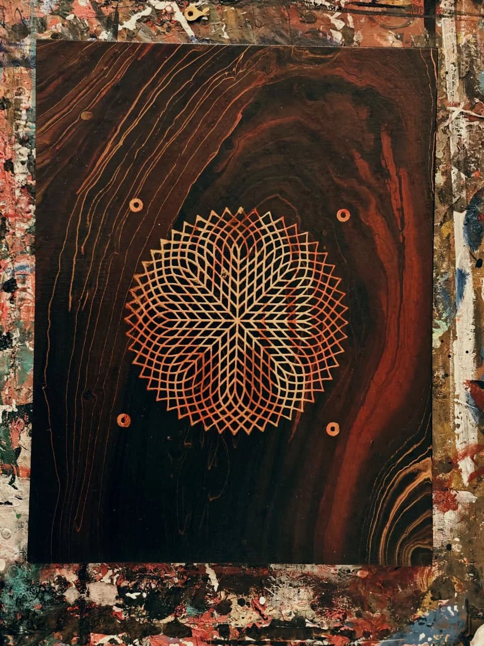 A lattice of fine gold lines crossing into an eight-petalled star at the centre of a dark red-brown panel painted with wood grain, four gold dots around it, on a paint-spattered studio floor.